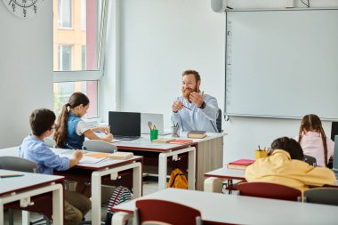 Bir erkek öğretmen, parlak, hareketli bir sınıf ortamında interaktif eğitimle meşgul olan bir grup öğrencinin önünde oturur..