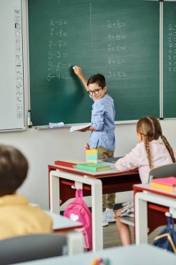 Çeşitli bir grup çocuk, parlak ve canlı bir sınıftaki erkek öğretmenlerinin talimatlarını dikkatle dinleyerek bir tahtanın önünde masalarında oturuyorlar..