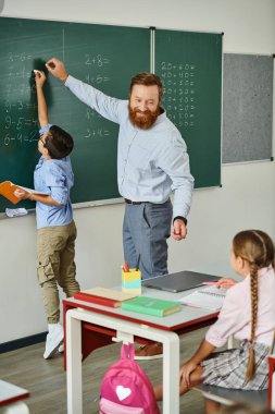 Bir erkek öğretmen, bir tahtanın önünde kendinden emin bir şekilde duruyor, parlak, canlı bir sınıfta bir grup çocuğu tutkuyla eğitiyor..