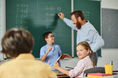 Bir grup farklı çocuk erkek öğretmenlerini dikkatle dinliyor parlak ve canlı bir sınıftaki bir tahtanın önündeki masada ders veriyorlar..