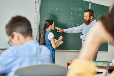 Kendini adamış bir öğretmen canlı bir sınıfın önünde duruyor ve bir grup çocuğa tahtanın önünde talimat veriyor..