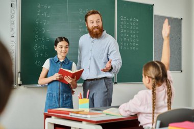 Bir adam tahtanın önünde küçük bir kızın yanında duruyor, canlı bir sınıfta ders veriyor..
