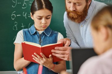 Genç bir kıza kitap okuyan bir erkek öğretmen hayat dolu bir sınıf ortamında ilgili bir ifadeyle.