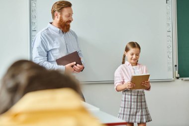 A man stands beside a little girl in front of a whiteboard in a vibrant classroom setting, engaging in a teaching moment. clipart