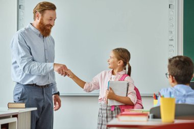 A male teacher in a vibrant classroom shakes hands with a young girl, fostering connection and respect between them. clipart