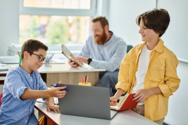 Masada oturan odaklanmış bir çocuk, canlı bir sınıftaki dizüstü bilgisayarı dikkatle inceliyor..