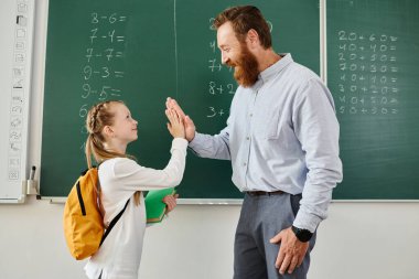 Bir adam ve küçük bir kız karatahtanın önünde duruyorlar, canlı bir sınıfta öğretmenlik yapıyorlar..