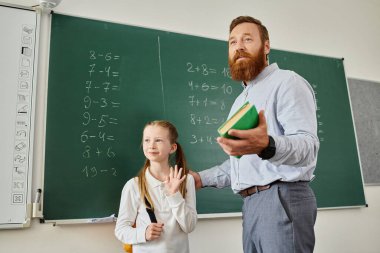 Sıradan giyinmiş bir adam küçük bir kızın yanında duruyor. İkisi de denklemler ve diyagramlarla dolu bir tahtaya dikkatle bakıyor..