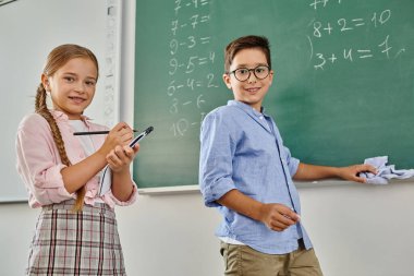 Oğlan ve kız, parlak ve canlı bir sınıfta öğrenmek için bir tahtanın önünde duruyorlar..