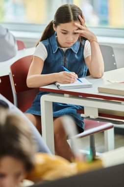 Genç bir kız elinde bir defter ve kalemle masada oturuyor. Kendini tamamen yazısına kaptırmış..