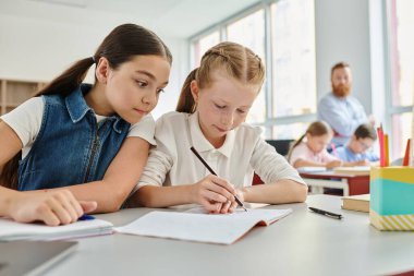 Two young girls are sitting at a table, immersed in their books, as they focus on reading and learning. clipart