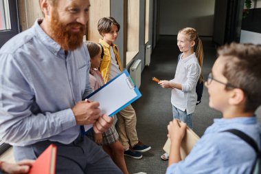 A male teacher holding a clipboard stands beside a group of kids in a vibrant classroom setting. clipart