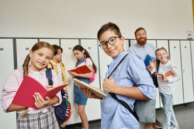 Erkek öğretmenlerinin rehberliğinde çeşitli bir grup çocuk parlak bir koridorda dikilip yeni bir öğrenme macerasına hazırlanıyorlar..