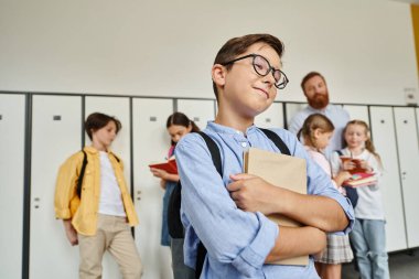 A young boy stands in front of a row of lockers, engrossed in a book he holds in his hands while in a school hallway. clipart