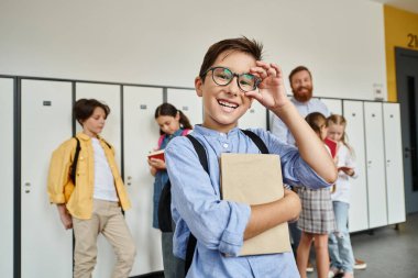 A boy in a blue shirt and glasses stands confidently in front of lockers in a school hallway. clipart