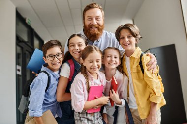 Bir erkek öğretmen, parlak bir eğitim ortamında bilgi ve rehberlik sunan çeşitli bir grup çocukla canlı bir koridorda karşılaşır..