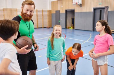 Bir erkek öğretmen basketbol topunu tutarken, bir grup çocuk onun etrafında parlak, canlı bir sınıfta dikiliyorlar..