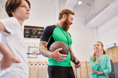 Basketbolu ustalıkla elinde tutan bir adam çocukların yanında duruyor, takım çalışması ve sportmenlik yayıyor..