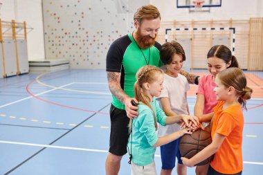 Basketbol sahasında çeşitli çocukların önünde duran bir adam onlara uygun teknik ve stratejiler öğretiyor..
