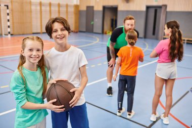 Bir grup çocuk, bir erkek öğretmen de dahil, bir spor salonunda basketbol aktiviteleri düzenliyorlar..