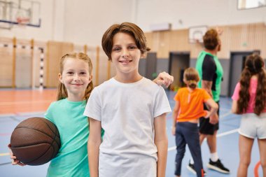 İki çocuk, bir erkek ve bir kız, yan yana dururlar, basketbol topunu tutarlar ve mutlu bir şekilde gülümserler..