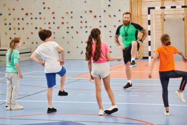 Çeşitli bir grup öğrenci, parlak bir sınıf ortamında öğretmenlerinden rehberlik alırken spor salonunda neşeyle duruyorlar..