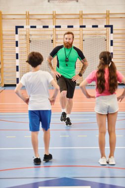 Bir erkek öğretmen tarafından yönetilen çeşitli bir grup çocuk, parlak bir spor salonunda birlikte egzersiz yapıyorlar..