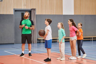 Bir adam elinde basketbol topuyla bir grup çocuğun önünde duruyor ve canlı, meşgul bir sınıf ortamında rehberlik ediyor..