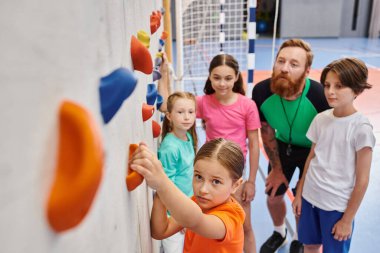 Bir grup genç ve çeşitli çocuk, erkek öğretmenlerinin onlara öğrettiği gibi parlak ve canlı bir spor salonunda yan yana duruyorlar..