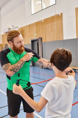 Yeşil tişörtlü bir adam ve beyaz tişörtlü bir çocuk parlak, canlı bir sınıf ortamında sohbet ediyorlar..