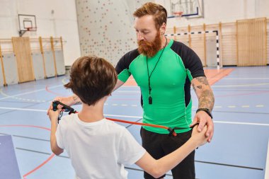Ayırt edici kızıl sakallı bir adam genç bir çocuğa canlı bir spor salonunda öğretiyor.