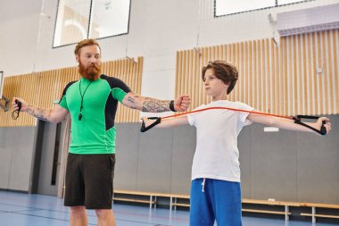 A bearded instructor with tattoos guides a young student through arm stretches in a vibrant school gymnasium. clipart