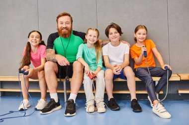 A man with a beard, a teacher, sitting on a bench surrounded by happy, diverse children of various ages, engaging in conversation and learning together. clipart