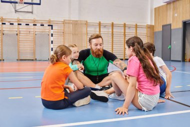 Bir erkek öğretmen, canlı bir okul spor salonunda, etkileşimli ve eğitici aktivitelerle meşgul çeşitli bir grup çocukla birlikte yerde oturuyor..