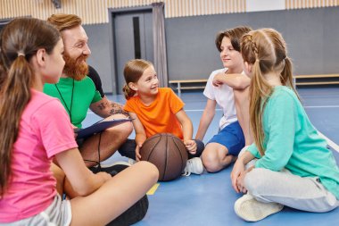 Çeşitli çocuklardan oluşan bir grup yerde oturup erkek bir öğretmenin talimatlarını dinliyorlar, ortada bir basketbol topu..