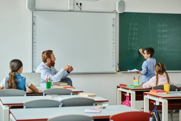 A group of children sits attentively at desks in front of a blackboard, listening to a male teacher who is instructing them in a bright and lively classroom setting.