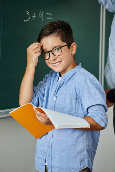 A young boy confidently stands in front of a blackboard, holding a book