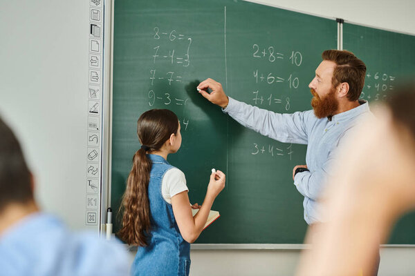 A man in colorful attire teaches a group of kids by a blackboard in a bright classroom setting.
