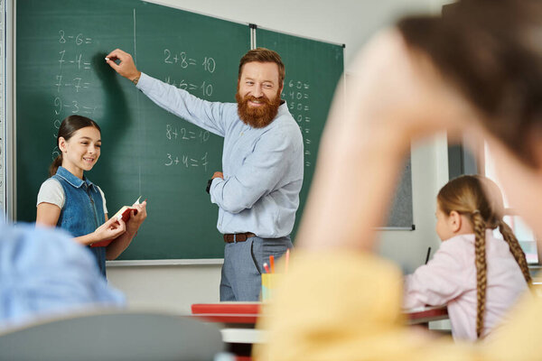 A male teacher stands before a blackboard in a vibrant classroom, instructing a group of children with enthusiasm and expertise.