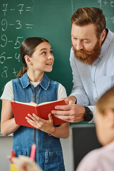 A man stands next to a little girl in front of a blackboard, engaging in an educational discussion in a vibrant classroom.