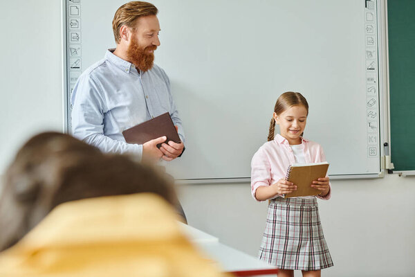 A man stands beside a little girl in front of a whiteboard in a vibrant classroom setting, engaging in a teaching moment.