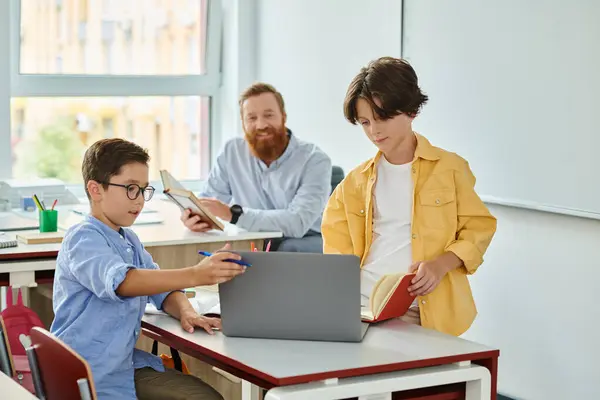 一个小男孩专心致志地坐在笔记本电脑前 而一个老师则在明亮活泼的教室里仔细观察着 — 图库照片