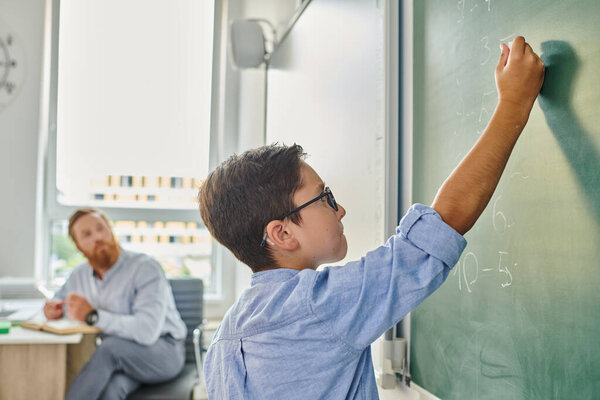 A young boy enthusiastically writes on a blackboard while a man teacher instructs him