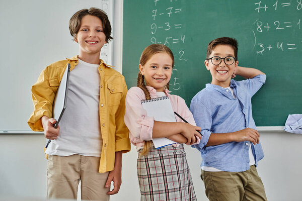 A group of children stand attentively in front of a chalkboard in a bright, lively classroom.