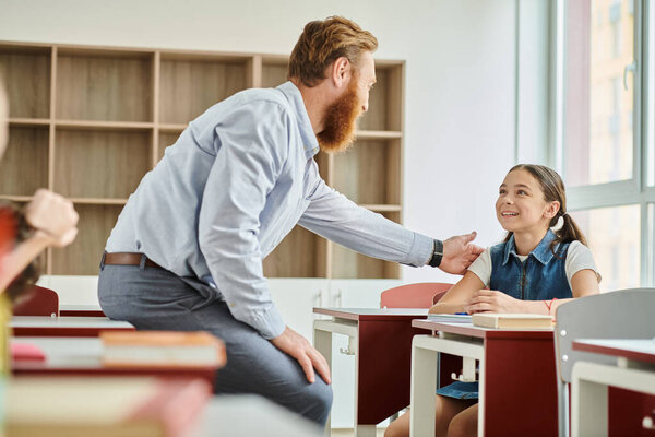 A man teacher encouraging little girl in a colorful, energetic classroom setting.
