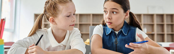 Two girls with pens in hand, focused and engaged in a creative task at a brightly lit classroom table.