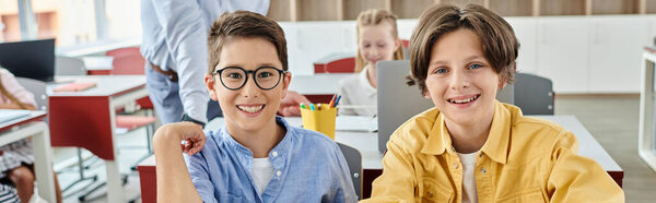 Два мальчика глубоко сосредоточены, сидят за столом в оживленном классе, пока их учитель инструктирует группу учеников.