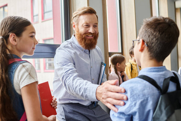 A diverse group of young people stand in a circle in a bright classroom, listening attentively to their male teachers instructions.