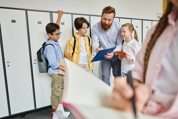 A man teacher is instructing a group of kids standing near lockers in a brightly lit classroom.
