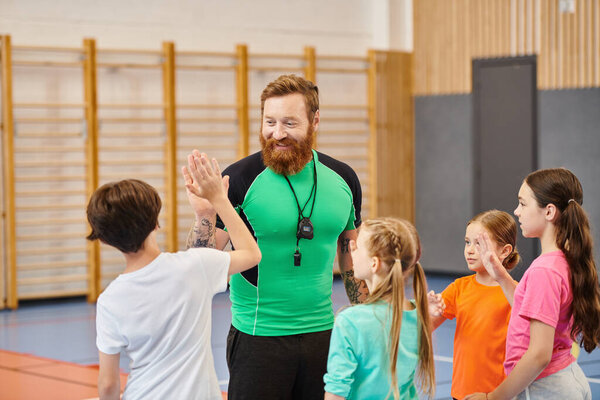 A bearded man stands confidently in front of a group of children, engaging them in a lively classroom setting.